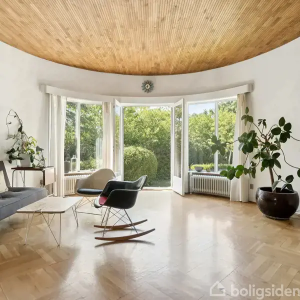 En stue med buet træloft, store vinduer og udsigt til frodig have. Sofa og gyngestol står ved vægkunst og planter. En rød "S" dekoration hænger på væggen.