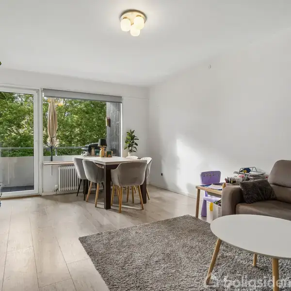 Stue med en brun sofa og rundt sofabord står på et tæppe. Spisebord med stole står foran en stor altandør med udsigt til grønne træer.