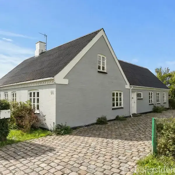 Hvidt køkken med beige flisegulv, der viser skabe og en køkkenmaskine på bordpladen. En buet døråbning fører til en gang med lyse vægge.