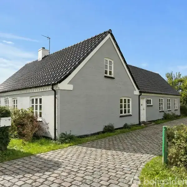 Gråt hus med sort tag står stille. Detaljerede hækplanter omgiver det på en brostensbelagt indkørsel i en stille forstadshave under en klar blå himmel.