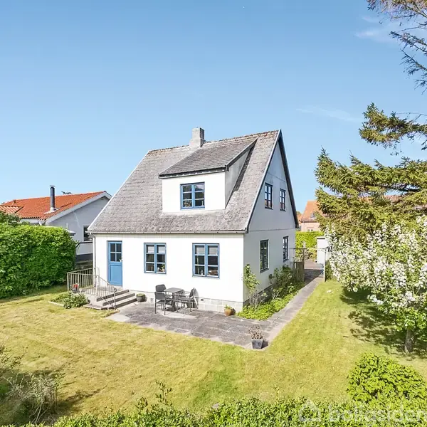 Et hvidt hus med blå vinduer står i en velplejet have. To havemøbler er placeret på en terrasse, omgivet af grønne buske og træer under en klar blå himmel.