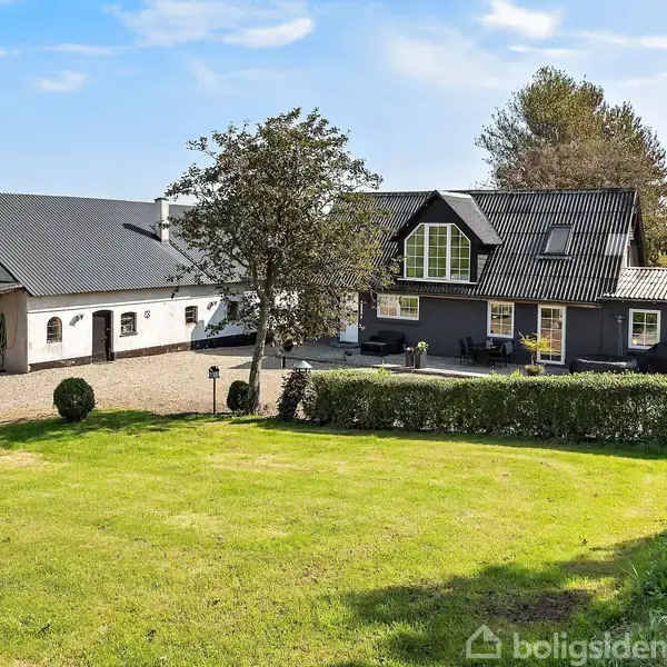 Et landhus med sort tag og lyse vægge står stille ved siden af en garagelignende bygning. Foran huset er en velplejet græsplæne, omgivet af buske og træer, under en blå himmel.