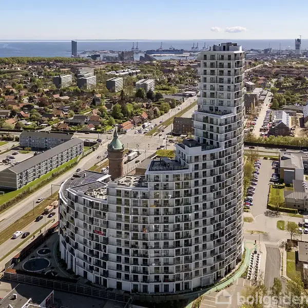 En stor, moderne bygning i buet form stiger op i et bybillede med beboelsesejendomme, en kirkeagtig struktur, og havnen i baggrunden.