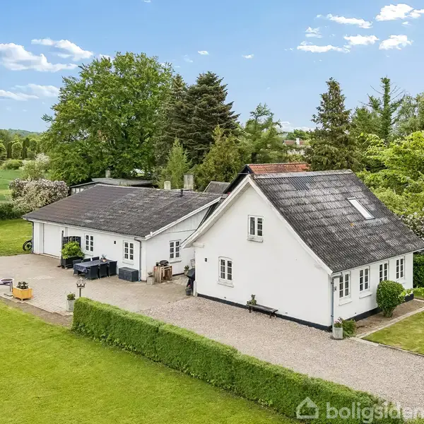 Et hvidt hus med sort tag står stille på en grusvej. Omkring det er en velplejet græsplæne og træer under en blå himmel med få skyer.