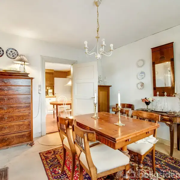 Et træspisebord står midt i en spisestue, omgivet af stole. Et skab med skuffer står til venstre, og et vægspejl hænger over et konsolbord. Rummet har en tæppebelagt gulv og en lysekrone.