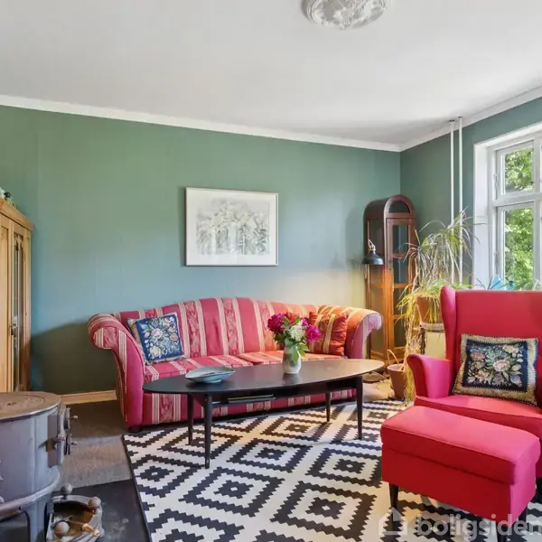 En stue med en rød sofa og lænestol, begge dekoreret med mønstrede puder. Et sort-hvidt tæppe dækker gulvet. Der er et brændeovn, træskab og grønne vægge.