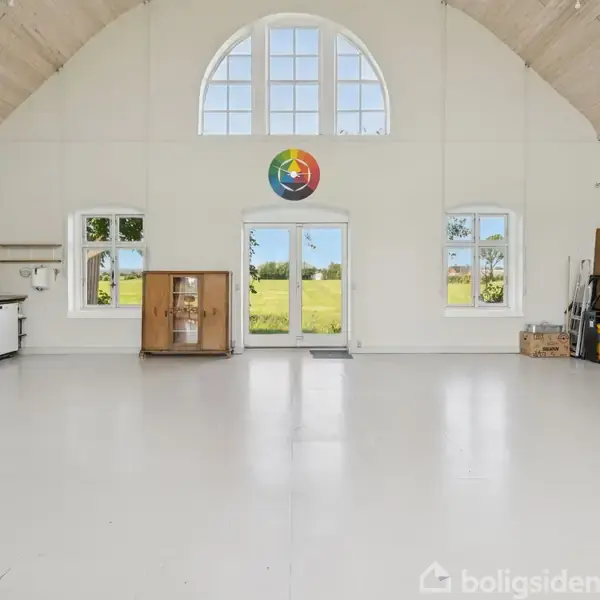 Rummeligt, lyst værelse med højt til loftet, store buede vinduer, og en dør i midten åbner til en grøn mark. Reol med bøger og diverse genstande langs højre væg.