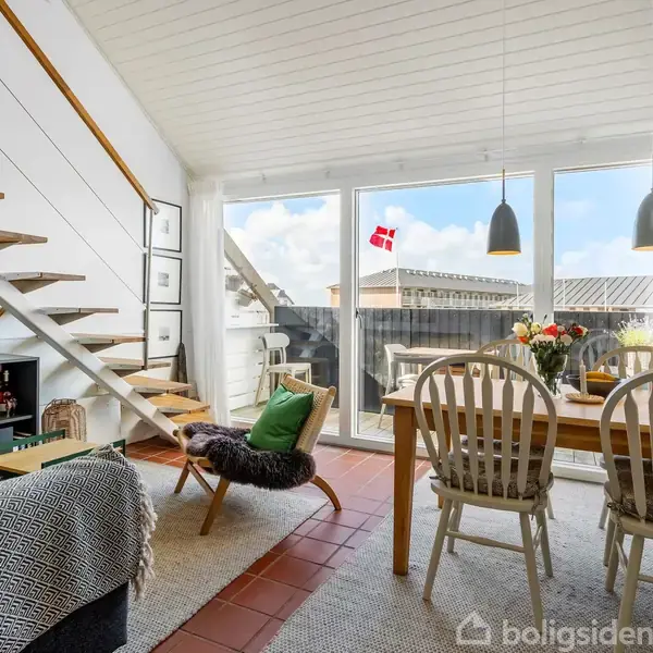 Stue med trappe til venstre, som fører opad. Til højre er et spisebord med blomster, omgivet af stole. Store vinduer bagved viser en balkon med et dansk flag.