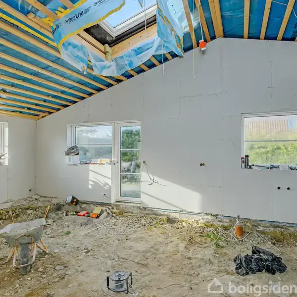 Et rum under renovering med ufærdigt gulv og synlige loftsbjælker. Værktøj og byggematerialer ligger rundt i rummet. Dagslys strømmer ind gennem to vinduer og en glasdør.