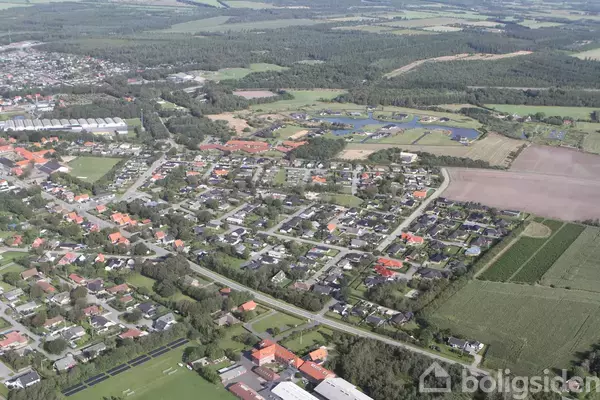 En by set fra luften med et netværk af veje og huse omgivet af marker og skove i baggrunden. Området ser fredeligt og landligt ud.