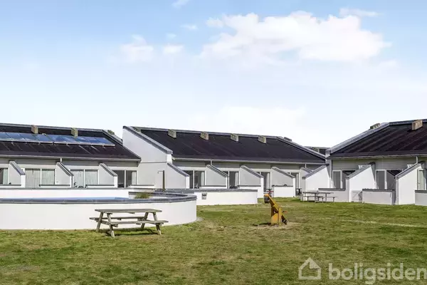 Rækkehuse med sorte tage står på række. Foran dem en bred græsplæne med to picnicborde og en lille legeplads. Himlen er blå med få skyer.