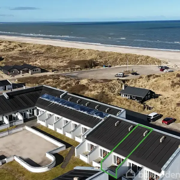 En kystnær bygning med sort tag, afslappet foran for en bred sandstrand og hav, omgivet af klitter og flere mindre huse. Parkerede biler ses nær bygningerne.