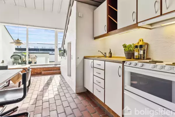 Et køkken med hvidt skab og komfur på højre side. Et bord med sorte stole til venstre. Store vinduer giver udsigt til en terrasse udenfor.