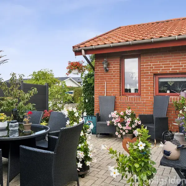 En hyggelig terrasse med sorte rattanmøbler omkring et bord, omgivet af blomsterkrukker. Rød murstenshus i baggrunden under en klar blå himmel, skaber en indbydende atmosfære.