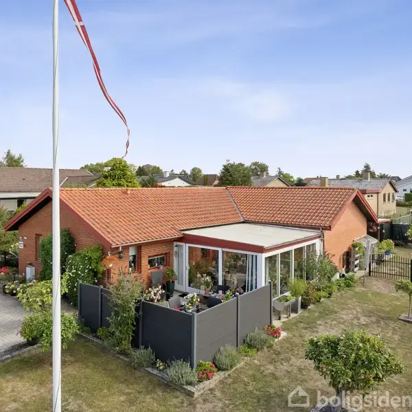 Et rødt murstenshus med rødt tegltag står stille midt i en velplejet have. Flagstang til venstre. Omkringliggende huskvarter set i baggrunden under en klar blå himmel.