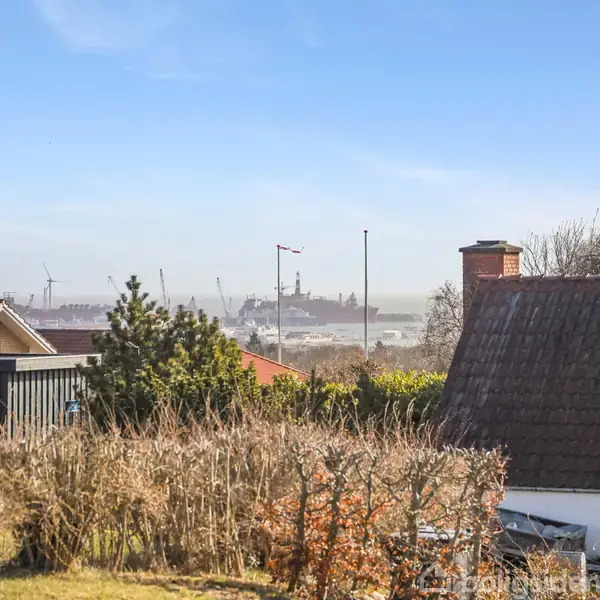 Et hus med skråt tag står stille omgivet af en have med buske. I baggrunden ses en by med skorstene og en færge i horisonten under en blå himmel.