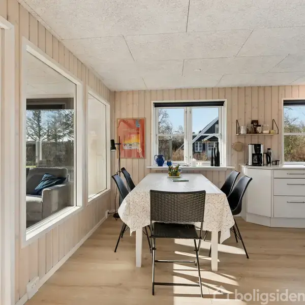 Spisestuebord med hvid dug og fire stole står i et lyst rum. Til venstre ses en stue med sofaer, og til højre er der et køkkenområde.