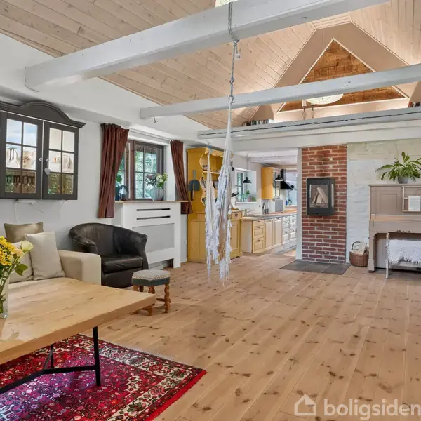 Stue med træmøbler, sofa, lænestol, og træbord med blomsterbuket på rød tæppe. Pejs midt i rummet. Køkken i baggrunden. Loft med hvide bjælker og skråtag. Indrettet med varme farver.