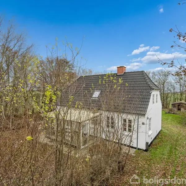 Et hvidt hus med gråt tag står omgivet af løvtræer og en grøn græsplæne. Huset har et vedhæftet drivhus. Himlen er blå med lette skyer.