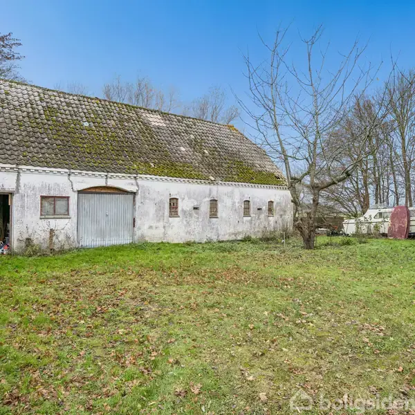Ældre hvid bygning med skråt tag og mos, står i en have med en stor græsplæne, omgivet af træer og en blå himmel i baggrunden.