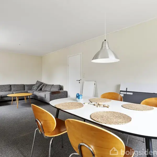 Spisebord med træstole er dækket med bordskånere, placeret i en stue med gråt tæppe. I hjørnet står en grå hjørnesofa og et lille sofabord ved hvide vægge.