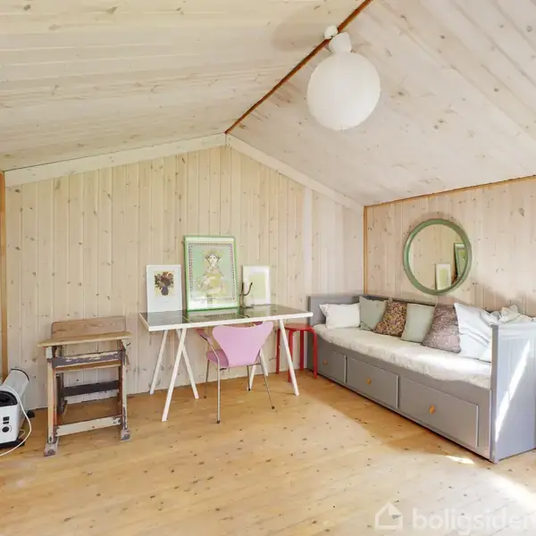 Et loftsrum med skrå vægge indeholder en grå daybed med puder, et skrivebord med stole og kunst på væggen. Rundt spejl og lille vindue giver naturligt lys.