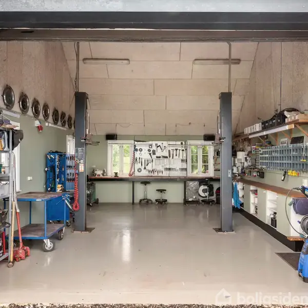 Garage med åbne døre og organiseret værkstedsområde. Værktøj hænger på væggene, borde og hylder står langs begge sider. Vinduer bagerst lukker lys ind og skaber en arbejdsstation.