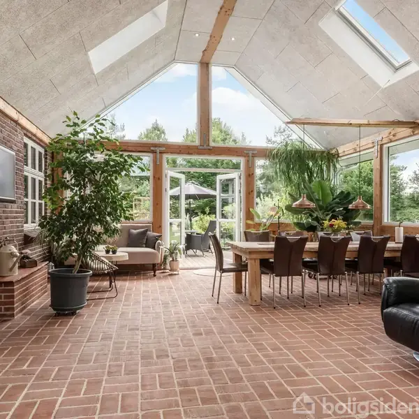 Plante i potte placeret i rummelig udestue med glasvægge. Langt spisebord omringet af stole i træmiljø. Udenfor vises grønne træer. Skylights på taget tillader naturligt lys.