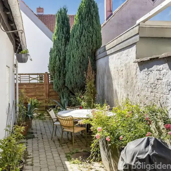 En terrasse med et bord og stole omgivet af blomsterbede og planter. Stedet er indrammet af hvide og grå mure, med nogle træer i baggrunden og klart sollys.
