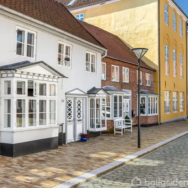 Række af charmerende byhuse står stille langs en brostensbelagt gade i solskinsvejr. Husene varierer i farver, herunder hvid og gul, med dekorative vinduer og en gadelampe foran.
