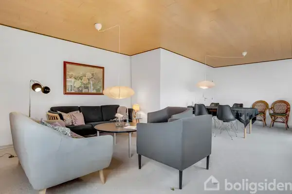 Gråt loungesæt med sofabord i en moderne stue. Billede på væggen og to pendellamper oplyser rummet, som har et stort spisebord omkranset af stole i baggrunden.