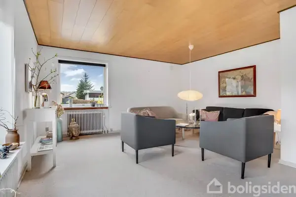 Stue med grå sofa og to stole står omkring et sofabord. En lampe hænger fra loftet. Vægdekorationer pryder rummets hvide vægge, og et vindue giver udsigt til træer udenfor.