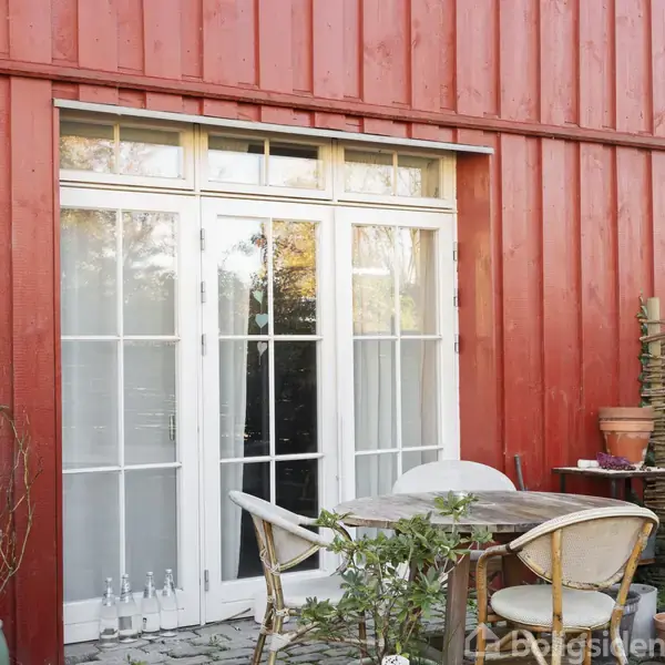 En hvid dør sætter i en rød trævæg. Foran døren står et lille bord og to stole i en terrasse omgivet af planter.