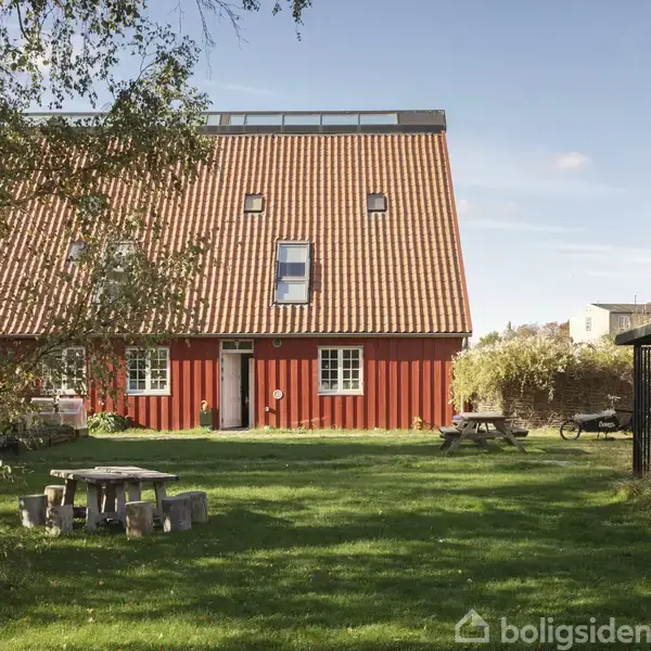 Rødt hus med skråt tegltag, står på græsplæne med lys fra solen. Tæt på er der træer, et picnicbord og nogle bygninger i baggrunden.