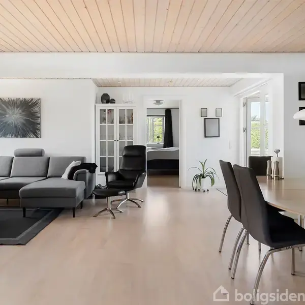 En stue med gråt sofahjørne og sort lænestol til venstre. Et spisebord med sorte stole står til højre. Trægulv og -loft skaber en lys og moderne atmosfære.