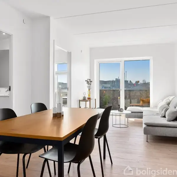 Spisebord med sorte stole står i et lyst rum. Sofa og sofabord i baggrunden nær store vinduer. Moderne, minimalistisk indretning med enkel dekoration og lys træfarvet gulv.