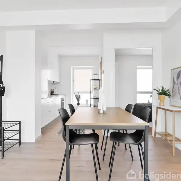 En træspisebord med sorte stole står centreret i en lys, moderne spisestue. Til venstre en sort reol, og bagved ses et hvidt køkken. Væggen til højre har en tegning og planter.