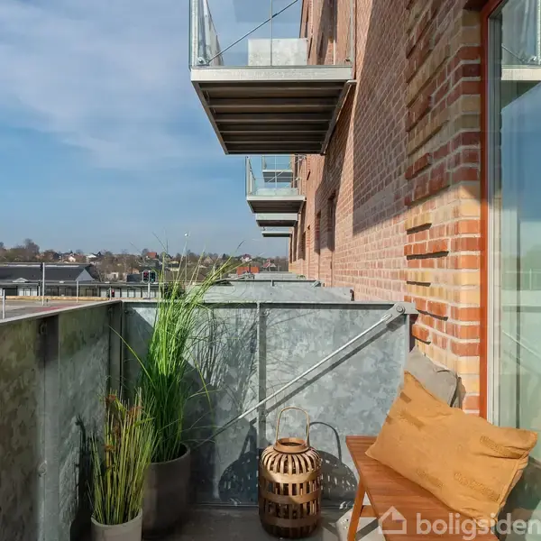 En altan i en murstensbygning er omgivet af glasværn. To dekorative puder ligger på en stol ved siden af en lanterne og en plante, der skaber en hyggelig atmosfære.