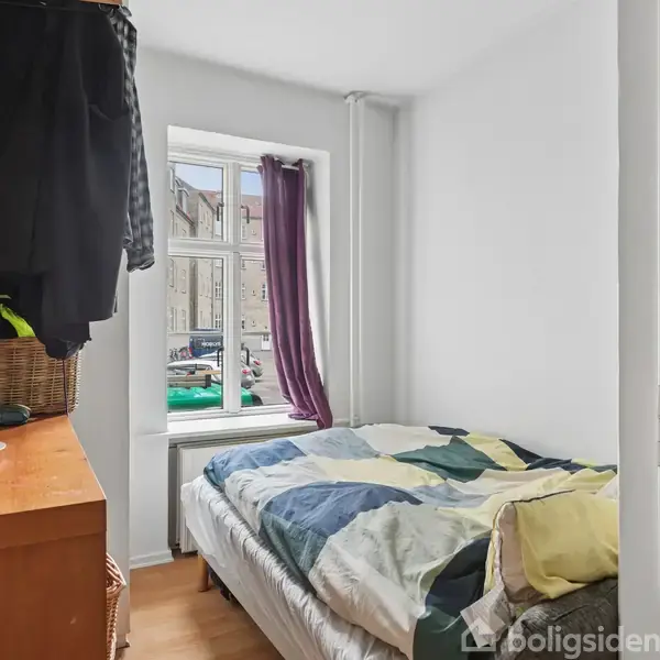 En seng med flerfarvet dyne står i et lille, lyst værelse. På kommoden ligger tøj, en højttaler og et tændt stearinlys. Et vindue med lilla gardin viser gadeudsigt.
