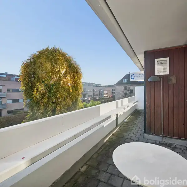 En rund hvidt bord står på en altan med flisebelægning, omgivet af en hvid træbalustrade og en brun væg med en lampe. Beboelsesbygninger og et træ ses i baggrunden.