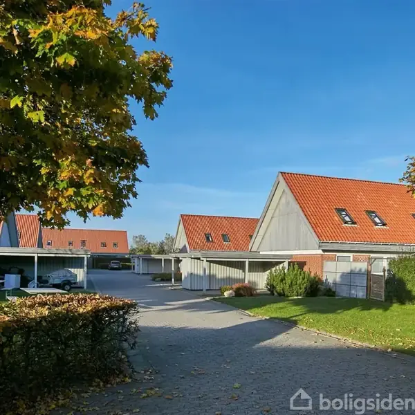 Rækkehuse med røde tegltage står stille i et solbeskinnet nabolag. Omkring dem er grønne træer og hække i et roligt, beboelsesområde med blå himmel.