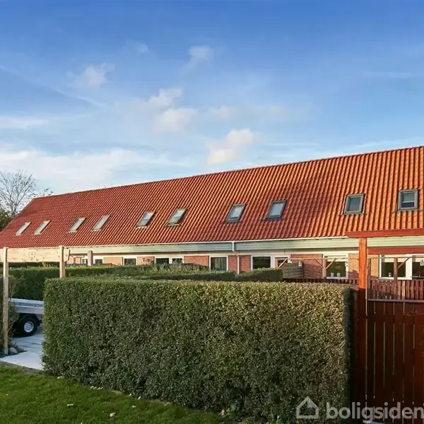 Rækkehus med rødt tag ligger bag en velplejet hæk og en træport; himlen er klar og blå med få skyer.