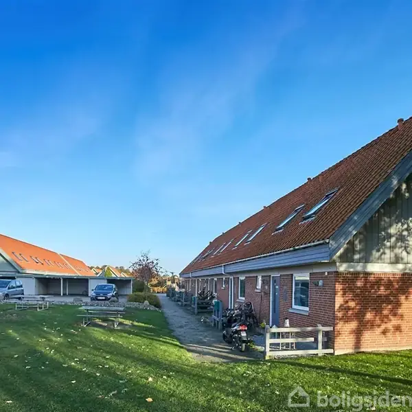 Rækkehus med rødt tag står stille under blå himmel, omgivet af græsplæne og parkeringsområde med biler i nærheden.