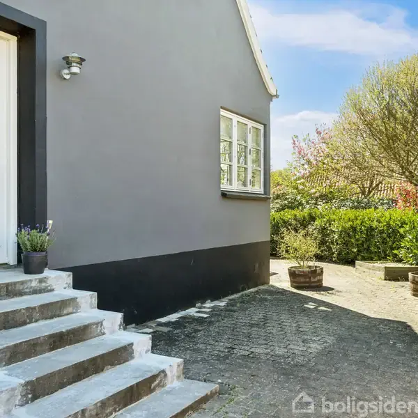En hvid dør med glasruder fører ind i et hus med grå facade. Trin med blomsterkrukker står foran døren. Omkring er en brostensbelagt gårdsplads med træer og buske.