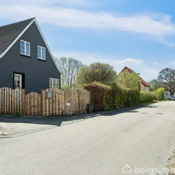 Et sortmalet hus står ved en stille vej, omgivet af en lav stakit og grønne hække på en solrig dag.