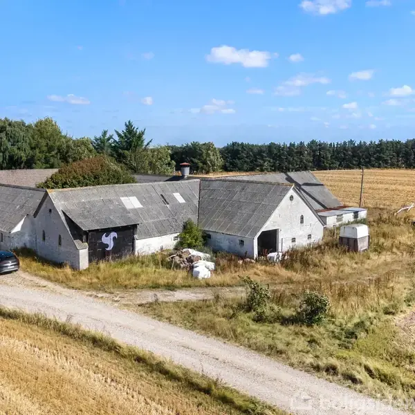 En gård med flere bygninger står stille i et landbrugsområde omgivet af gyldne marker. En vindmølle står ved siden af, og en bil er parkeret på grusvejen foran.
