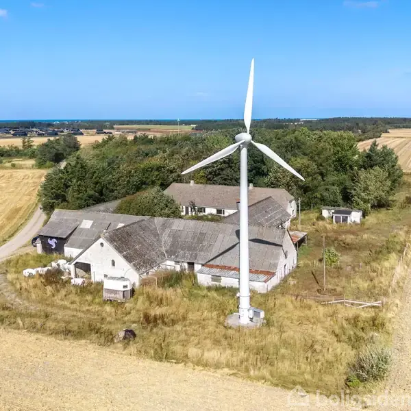 Vindmølle roterer ved siden af en ældre gård med flere bygninger. Omgivet af gyldne marker og en lille skov, og horisonten viser en blå himmel.