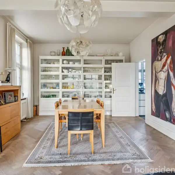Et spisebord med stole står centralt i et elegant rum med hvidt skab fuld af porcelæn. Store kunstværker hænger på væggene, og der er en moderne lysekrone i loftet.