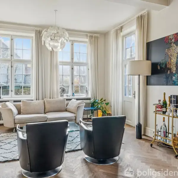 En lys stue med to sorte lænestole foran en cremefarvet sofa. En moderne lysekrone hænger fra loftet. Ved siden af sofaen står en barvogn med flasker.