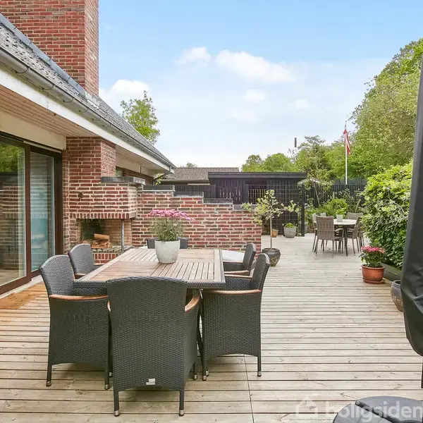Et udendørs terrasseområde med et bord og flere stole lavet af mørkt flet. Terrassen er omgivet af røde murstensvægge og hække, med udsigt til en grøn have.